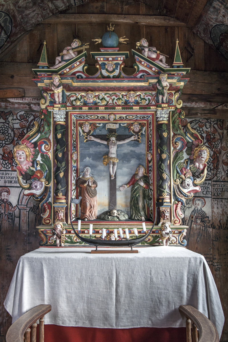 Utskåret Kristus på korset og Maria og Johannes. Under korset stener med hodeskalle og ben. Ved hver hånd englehode med vinge. Bakgrunn malt med grå skyer og gul solnedgang og byprospekt. Figurene lyst hudfarget, korset mørkt brunt. Kristi klede gult. INRI i gull på hvit bunn. Maria med rosa kjortel og brun kappe med gull, hvit hodelin. Johannes i rød kjortel og grønn kappe. Rundt tavlen stilisert ornamentikk i gult, grønt, brunt på hvit bunn. Englehoder midt på hver side. På begge sider vridde brune søyler med drueranker i grønt og gull, sokkel med hode, og kapitel øverst. Under det hele en sokkel med bladverk i gull og rødt på hvit bunn.