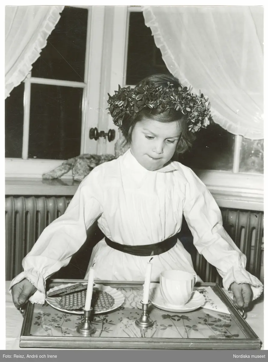 En flicka klädd i Luciaklänning, med en krans på huvudet, ställer en dukad bricka på bordet till Luciakaffet.