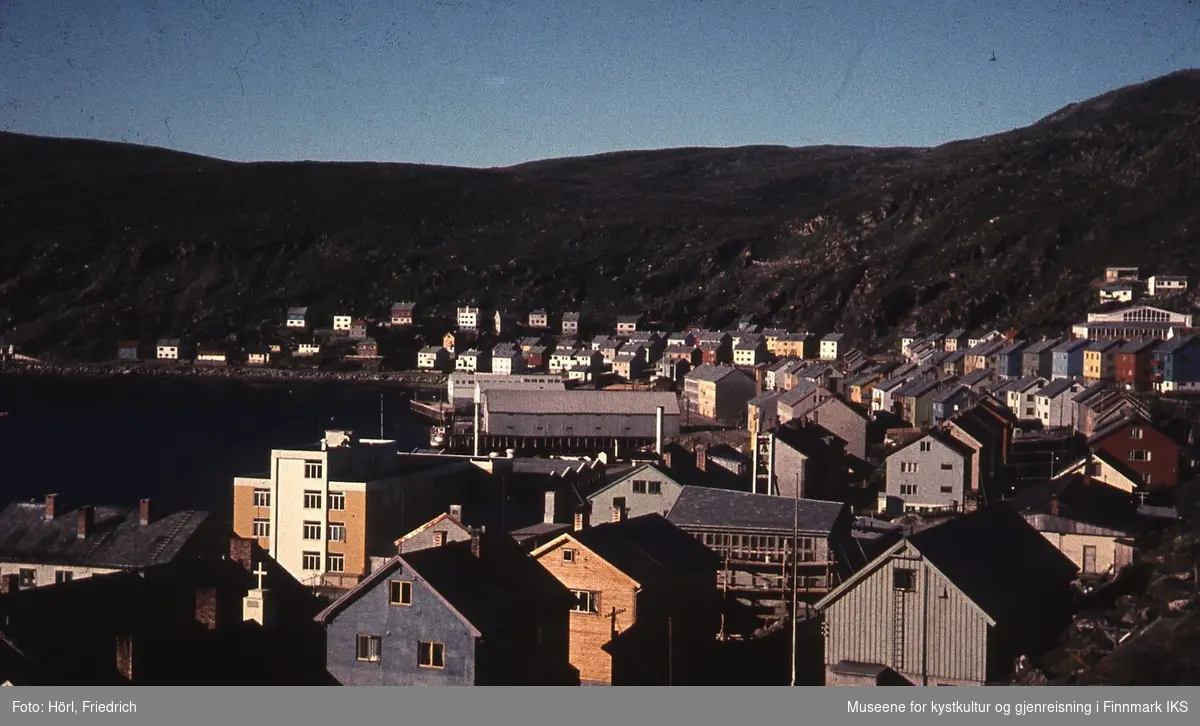 Utsikt over Hammerfest sentrum, bydelen Molla og Molla fjellet i bakgrunnen. Havna til venstre. Sola skinner. Mye av byen er gjenreist i den karakteristiske arkitekturen. Markante bygninger er Findus fabrikken nede til venstre, den katolske kirken i byggefasen og lagerhusene til Hauans materialhandel midt i bildet.