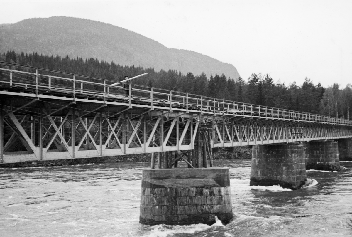 Reparasjon av jernbanebrua over Lågen nedenfor Hunderfossen i Øyer i Oppland. Fotografiet er tatt i slutten av mai 1940, en snau måned etter at tyske militære invaderte Gudbrandsdalen. De norske militære, som forsøkte å stanse eller bremse okkupasjonsstyrkenes framrykking, blant annet ved å ødelegge veg- og jernbanebruer. På Dovrebanen gjennom Gudbrandsdalen gikk det ut over Hunderbrua, Randkleivbrua i Ringebu og Ottabrua i Sel. Etter at okupantene fikk kontroll ble det en prioritert oppgave å få kommunikasjonsårene i stand igjen. Vi vet foresløpig ikke hva skadene på Hunderbrua besto i, men den skal ha blitt reparert i løpet av andre halvdel av mai 1940. Dette bildet er tatt i slutten av denne prosessen. Oppe på brua ser vi en arbeider med en planke på skuldra.