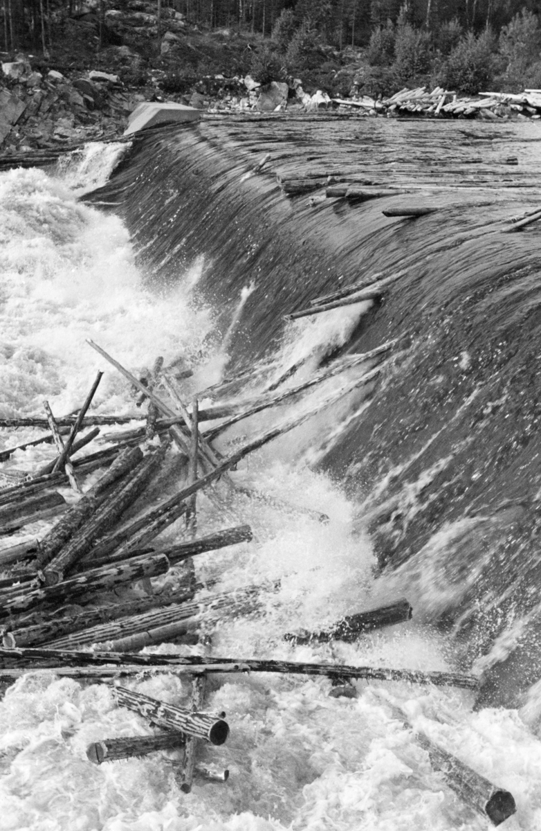 Dammen som skulle stuve opp vann til Kvernfallet kraftverk i elva Søndre Osa i Åmot kommune i Hedmark, fotografert i juni 1940. Her renner vannet over en avrundet terskel, utført i armert betong. Denne konstruksjonen er plassert på tvers av elveløpet, like framfor ei oppoverskrånende bergflate, som leder vannet fra nedfallssona mot den søndre elvebredden, før det føres videre vestover i det gamle elveløpet. Da dette fotografiet ble tatt gikk det tømmer i elva, og det var tilsynelatende i ferd med å bygge seg opp en tømmerhaug i den kvitskummende fallsona under dammen.