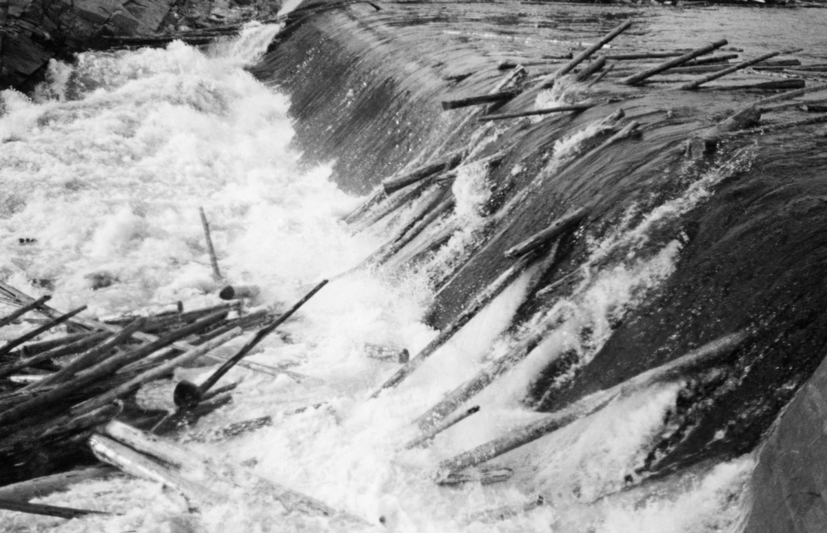 Dammen som skulle stuve opp vann til Kvernfallet kraftverk i elva Søndre Osa i Åmot kommune i Hedmark, fotografert i juni 1940. Her renner vannet over en avrundet terskel, utført i armert betong. Denne konstruksjonen er plassert på tvers av elveløpet, like framfor ei oppoverskrånende bergflate, som leder vannet fra nedfallssona mot den søndre elvebredden, før det føres videre vestover i det gamle elveløpet. Da dette fotografiet ble tatt gikk det tømmer i elva, og det var tilsynelatende i ferd med å bygge seg opp en tømmerhaug i den kvitskummende fallsona under dammen.