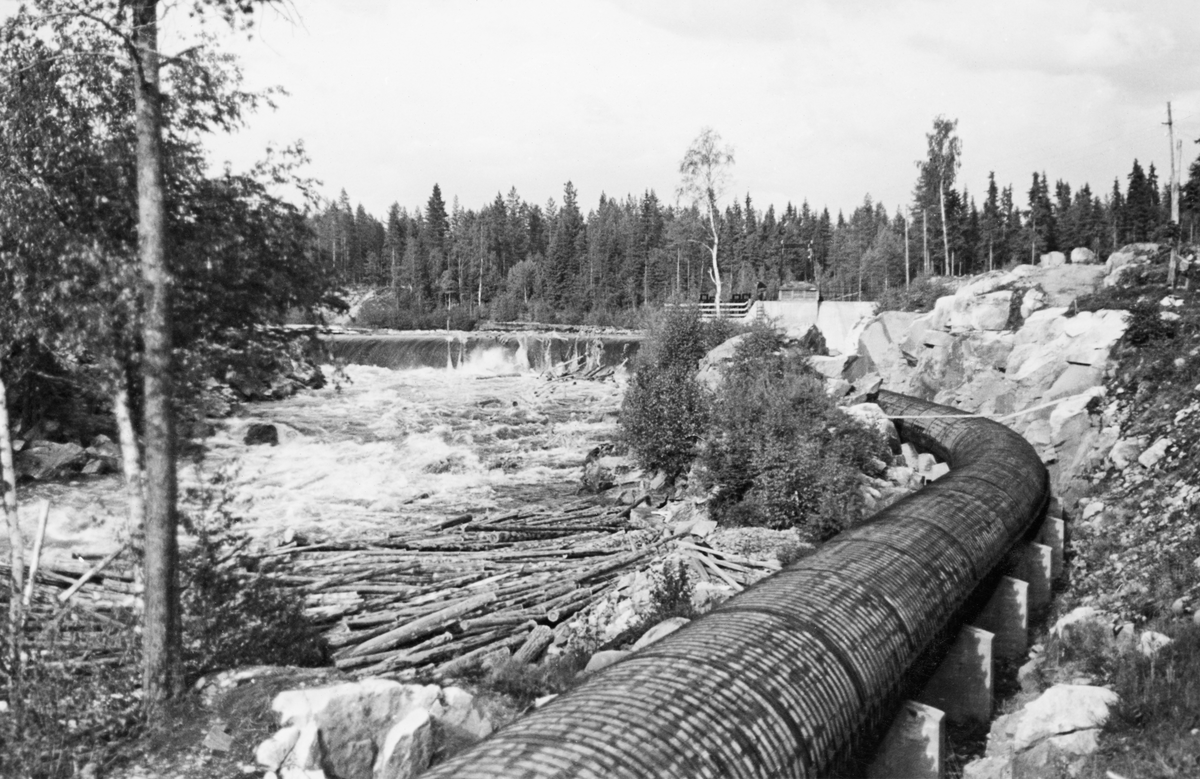 Dammen som skulle stuve opp vann til Kvernfallet kraftverk i elva Søndre Osa i Åmot kommune i Hedmark, fotografert i juni 1940. Her renner vannet over en avrundet terskel, utført i armert betong. Denne konstruksjonen er plassert på tvers av elveløpet, like framfor ei oppoverskrånende bergflate, som leder vannet fra nedfallssona mot den søndre elvebredden, før det føres videre vestover i det gamle elveløpet. I forgrunnen og til høyre i bildet ser vi turbinrøret, som skulle lede vann fra et basseng i enden av nedover mot kraftstasjonen ved Brufallet. Røret hadde et rundt tverrsnitt og var lagd av bord som ble holdt ihop av tettsittende jerngjorder. Det kvilte på betongfundamenter i en forsenkning som stedvis var nedsprengt i berget på sørøstre side av elveløpet.