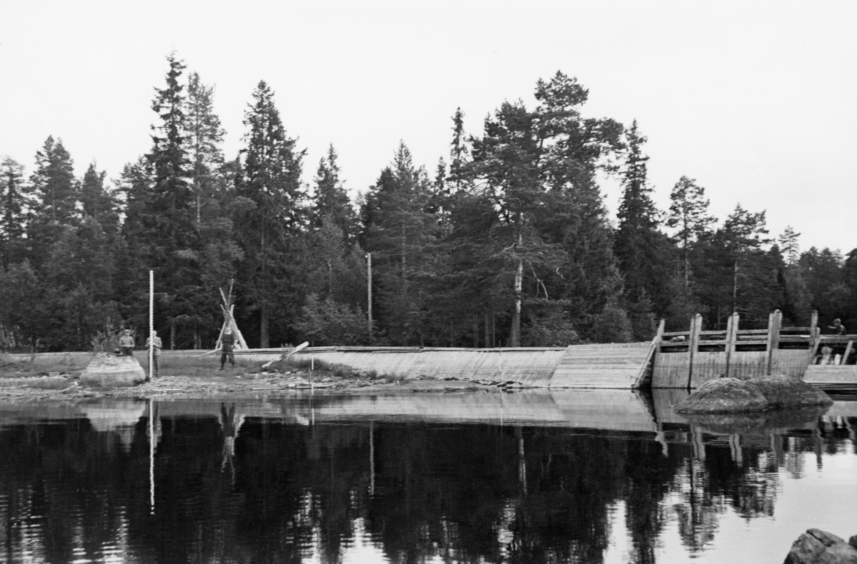 Fra Skasdammen på Brandval Finnskog, ved Skasåas utløp fra innsjøen Skasen. Fotografiet skal være tatt sommeren eller høsten 1940. Det viser en tredam, der sidekarene var konstruert som steinfylte tømmerkister. Disse konstruksjonene var imidlertid skjermet med et pulttak på oversida og med en skråvegg mot sjøen som skulle brukes som vannreservoar for fløtinga nedover i Skasåa. Både taket og veggskjermen var lagd av kavler. Damkaret på motsatt side av elveløpet ser ut til å være konstruert på samme måte. Over løpet gikk det ei trebru, og på damsida av den ser vi fire damluker, som ble brukt som stengsel når det skulle samles vann og som ble hevet når det ble sluppet vann for å få tømmeret til å flyte nedover i åa.