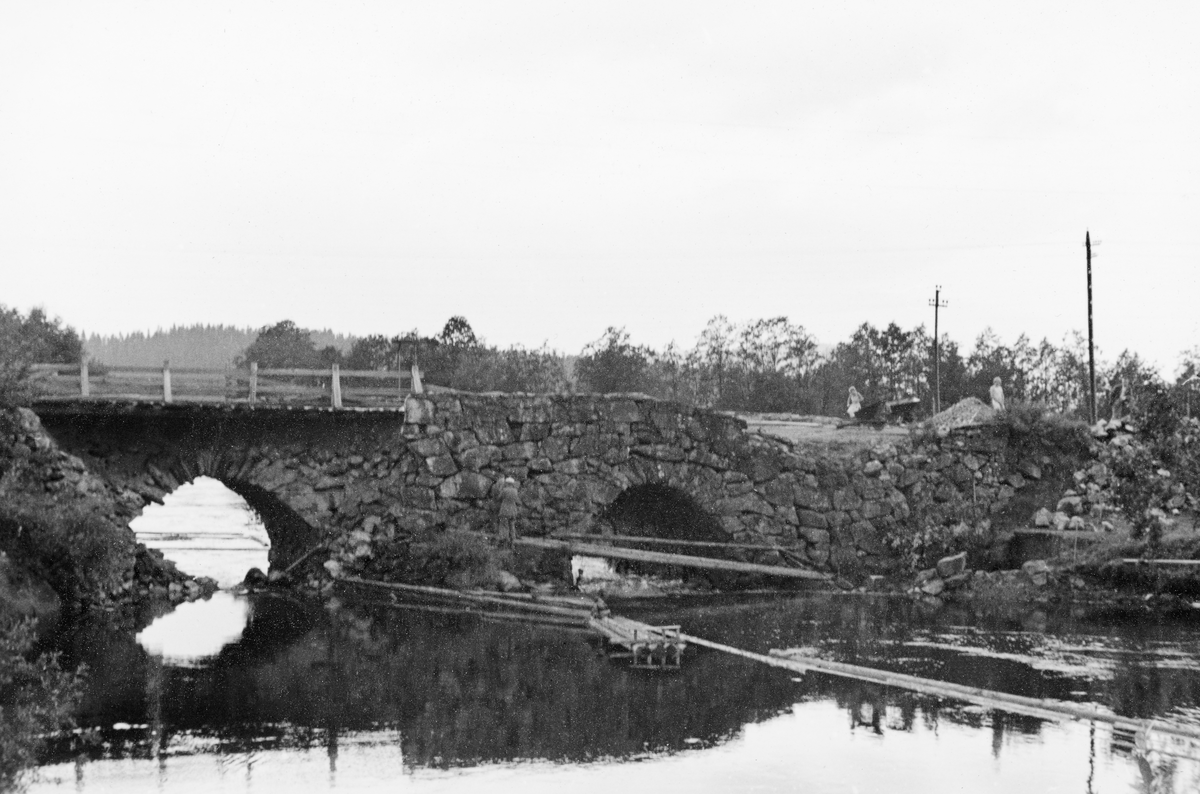 Fra reparasjonen av Norsbrua i Vinger i Hedmark, der vegen krysser elva fra Vingersjøen mot Glomma. Fotografiet ble tatt i juli 1940. Dette var ei natursteinsmurt hvelvbru med to løp. På dette fotografiet ser vi at det ene bruhvelvet er delvis istykkersprengt, For å gjøre brua farbar for kjøretøyer hadde man lagt en trebane oppå det gjenværende murverket, antakelig som et midlertidig tiltak. Ifølge avisoppslag i 1940 ble det ene hvelvet sprengt for å hindre de tyske styrkenes framrykking. Denne skaden ble reparert. Seinere på sommeren, i juli, da tømmeret fra Skasvassdraget kom, oppsto det en ny kritisk situasjon, og det endte med at man valgte å sprenge det andre bruhvelvet. Vegmyndighetene mente imidlertid at de skulle få brua istand igjen.