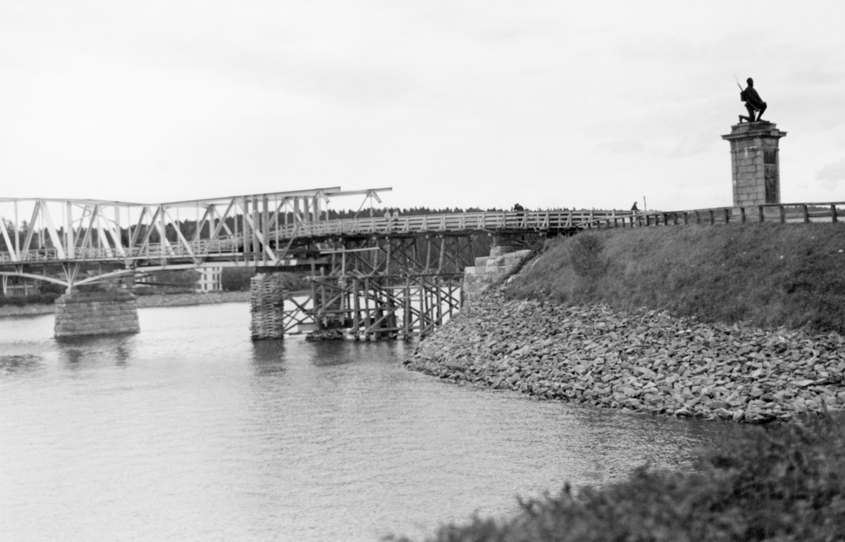 Brua over elva Vorma i Sundet - kommunesenteret i Eidsvoll på Øvre Romerike. Dette fotografiet ble tatt fra den vestre elvebredden, i medstrøms retning, fra en posisjon like ved jernbanestasjonen. Bildet viser hvordan norske styrker hadde sprengt det vestre bruspennet da de trakk seg tilbake på østsida av elva under invasjonen i 1940. På dette fotografiet, som ble tatt i slutten av august samme år, ser vi hvordan man, som et midlertidig tiltak, hadde bygd ei trebru som skulle gjøre det mulig for lette trafikanter å ta seg fra vestre elvebredd over til den gjestående delen av fagverkskonstruksjonen av jern. Sundbrua ble ferdigstilt i 1923. Mer informasjon om denne brua finnens under fanen «Opplysninger». Ved bruhodet ser vi Gustav Lærums (1870-1938) skulptur til minne om riksforsamlinga på Eidsvoll i 1814, en kneldende soldat, vendt mot Eidsvoll kirke. Skulpturen ble avduket i 1928.