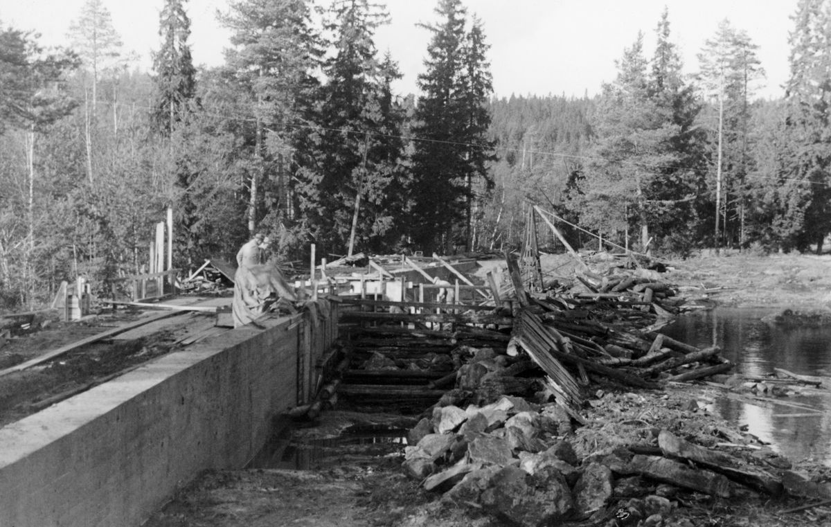 Bygging av ny dam, Skasdammen på Brandval Finnskog, ved Skasåas utløp fra innsjøen Skasen. Fotografiet skal være tatt høsten 1940. Bildet viser et anleggsområde der det var støpt nye betongmurer, både mot innsjøen Skasen, og i løpet der Skasåa rant ut av sjøen, hvor også tømmeret fra omkringliggende skoger skulle følge vannet. Da dette fotografiet ble tatt var et par karer - antakelig representanter for fløtingsadministrasjonen - på befaring. På motsatt side av det nevnte løpet ser vi ei kran.

Litt mer informasjon om vassdraget og Skasdammen finnes under fanen «Opplysninger».