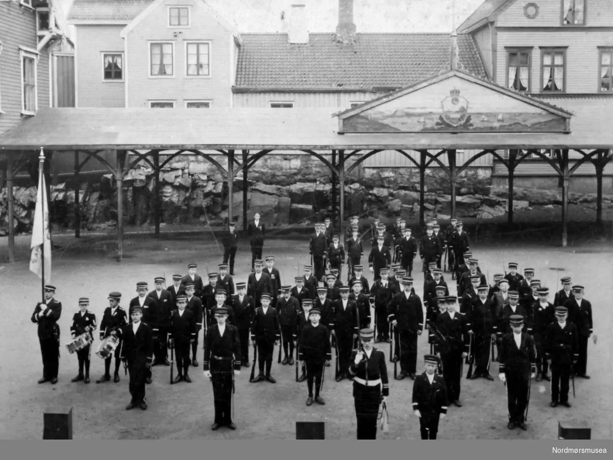 Reprofoto. Musikkorps oppstilt på skoleplana ved Enggatens pikeskole på Kirklandet i Kristiansund. Datering er ukjent, men muligens ved århundreskiftet (1890-1900). Bestillingen er registrert på Romsdalsposten. Reprofotograf er Nils Williams.