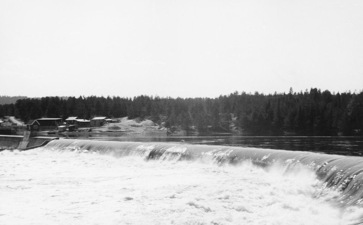 Dammen ved Skjefstadfossen i Elverum, fotografert fra den østre elvebredden med kameraet vendt mot den vestre sida av elva. Denne dammen ble bygd i 1909-1910 med sikte på at den skulle stuve opp vann til det nye kraftverket ved den vestre elvebredden i det ovenforliggende elveløpet. Dammen ble 312 meter lang, og den fikk en noe buet form. Damkrona ble støpt slik at den skrånet en aning nedover mot den østre enden. På den måten ønsket utbyggerne å trekke fløtingstømmeret mot den østre delen av elveløpet, for i vest lå kraftstasjonen og i midten av elva var det store steinører i sona nedenfor dammen, der tømmeret hadde lett for å sette seg fast.

Skjefstadfossen hadde opprinnelig en fallhøyde på sju meter.  Ved bygginga av kraftverksdammen som dette fotografiet er tatt fra i 1909-1910 ble vannspeilet ovenfor Skjefstadfossen noe hevet, slik at fallhøyden ble økt til 10,5 meter.  Dette anlegget var det første store kraftverket som ble reist i den øvre delen av Glomma (nord for innsjøen Øyeren).