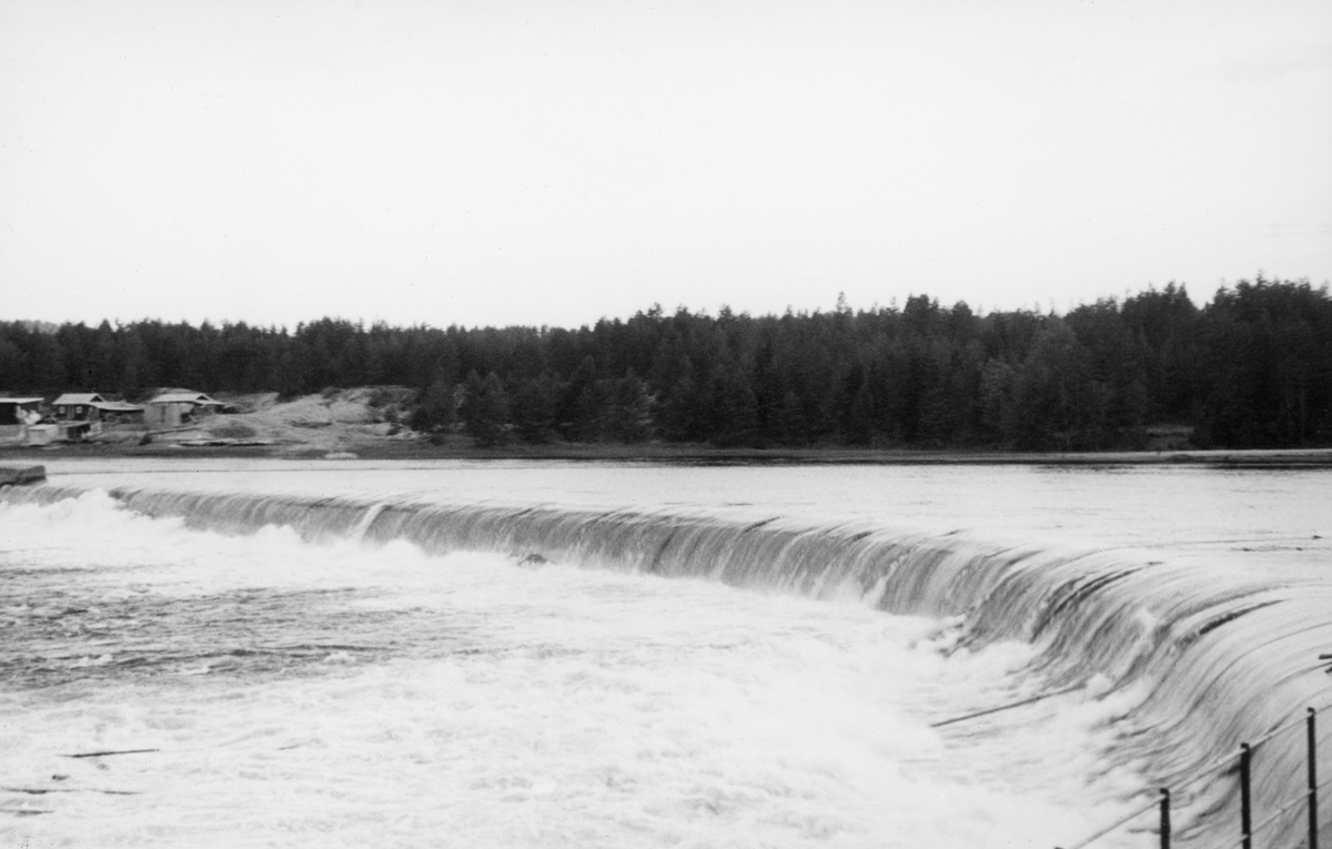 Dammen ved Skjefstadfossen i Elverum, fotografert fra den østre elvebredden med kameraet vendt mot den vestre sida av elva. Denne dammen ble bygd i 1909-1910 med sikte på at den skulle stuve opp vann til det nye kraftverket ved den vestre elvebredden i det ovenforliggende elveløpet. Dammen ble 312 meter lang, og den fikk en noe buet form. Damkrona ble støpt slik at den skrånet en aning nedover mot den østre enden. På den måten ønsket utbyggerne å trekke fløtingstømmeret mot den østre delen av elveløpet, for i vest lå kraftstasjonen og i midten av elva var det store steinører i sona nedenfor dammen, der tømmeret hadde lett for å sette seg fast. Da dette bildet ble tatt, i begynnelsen av juni 1941, var en del stokker på veg over damkrona. Bildet er tatt fra en plattform ved den østre elvebredden.

Skjefstadfossen hadde opprinnelig en fallhøyde på sju meter.  Ved bygginga av kraftverksdammen som dette fotografiet er tatt fra i 1909-1910 ble vannspeilet ovenfor Skjefstadfossen noe hevet, slik at fallhøyden ble økt til 10,5 meter.  Dette anlegget var det første store kraftverket som ble reist i den øvre delen av Glomma (nord for innsjøen Øyeren).