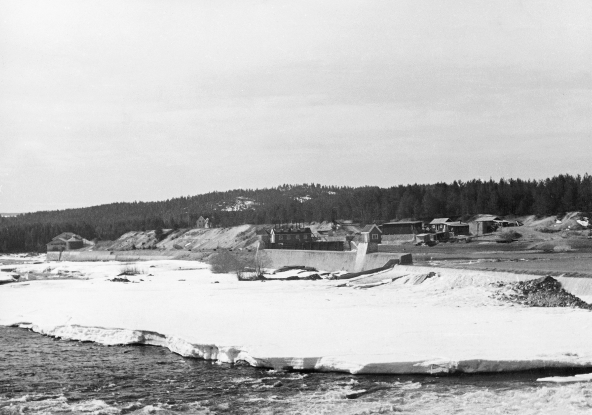 Kraftverksdammen ved Skjefstadfossen i Heradsbygda i Elverum, Hedmark, fotografert 1. mai i 1941. Fotografiet er tatt fra en posisjon på østre elvebredd, like nedenfor dammen. Dammen var 312 meter lang og hadde en buet profil. Den østre delen av dammen var noe lavere enn den vestre. Poenget med dette var å skape et «sug» som trakk tømmeret mot den østre delen av elveløpet, der elveløpet var djupest. Der var faren for at tømmeret skulle sette seg fast på elvebotnen og filtre seg sammen i hauger noe mindre enn i det vestre løpet. I den vestre enden av dammen var det et basseng med utløp for et grovt, om lag 400 meter langt turbinrør av betong som førte ned til kraftstasjonen, som vi ser til venstre i dette bildet. Våren var sein i 1941 - det lå fortsatt is i elveløpet like nedenfor den midtre delen av dammen.