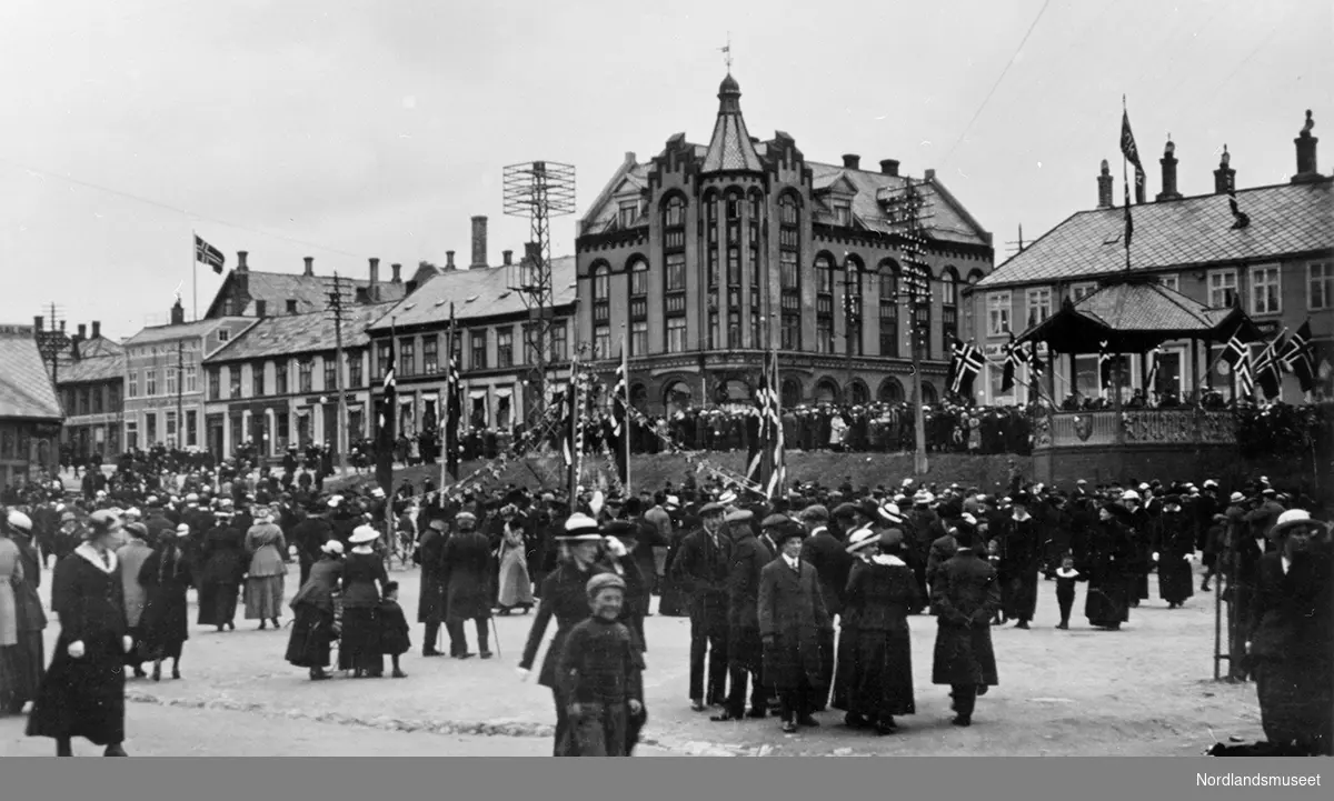 Postkort. Bodø torg, antagelig 17. mai. Folkemengde. Paviljongen til høyre. Bebyggelsen bak.