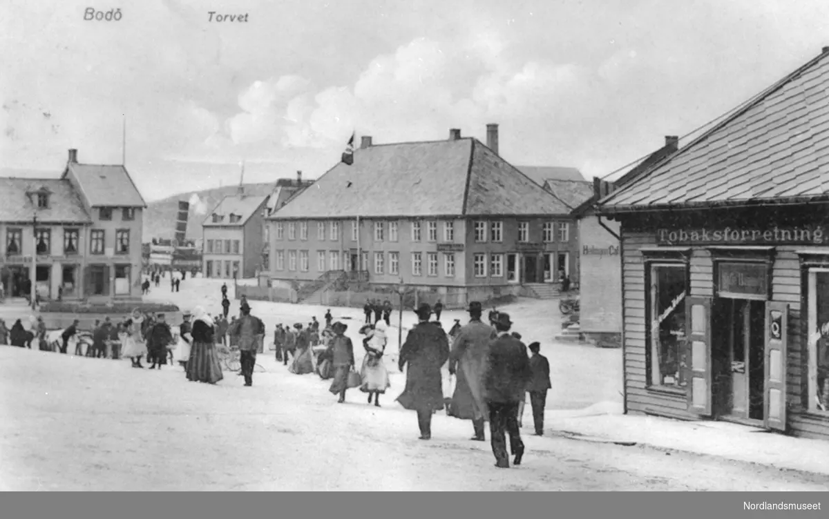 Postkort. Bodø, Torvet. Skråning ned mot bebyggelsen bak. Skorsteinen til en dampbåt mellom bygningene. Åpen plass foran med flere mennesker.