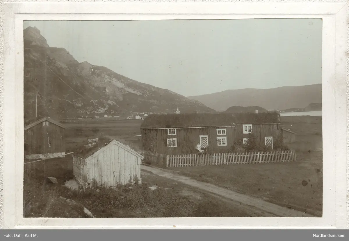 Kaffeselskap foran huset under Steigberget med stakittgjerde rundt og to uthus? på andre siden av veien, Steigen med kirken i bakgrunnen