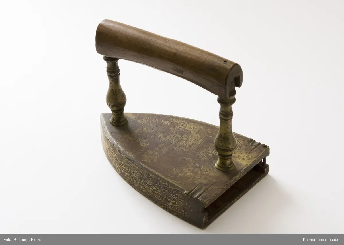 KLM 7067. Strykjärn. Tillverkat av malm. Vanlig form, framtill spetsigt, baktill tvärt avskuret. Översidan graverad med ett stort rosornament. Raka profilerade ståndare av malm. Hantag av trä. Luckan för lodet saknas.