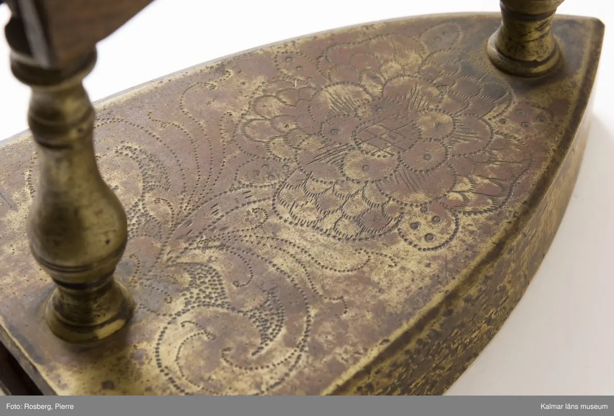 KLM 7067. Strykjärn. Tillverkat av malm. Vanlig form, framtill spetsigt, baktill tvärt avskuret. Översidan graverad med ett stort rosornament. Raka profilerade ståndare av malm. Hantag av trä. Luckan för lodet saknas.