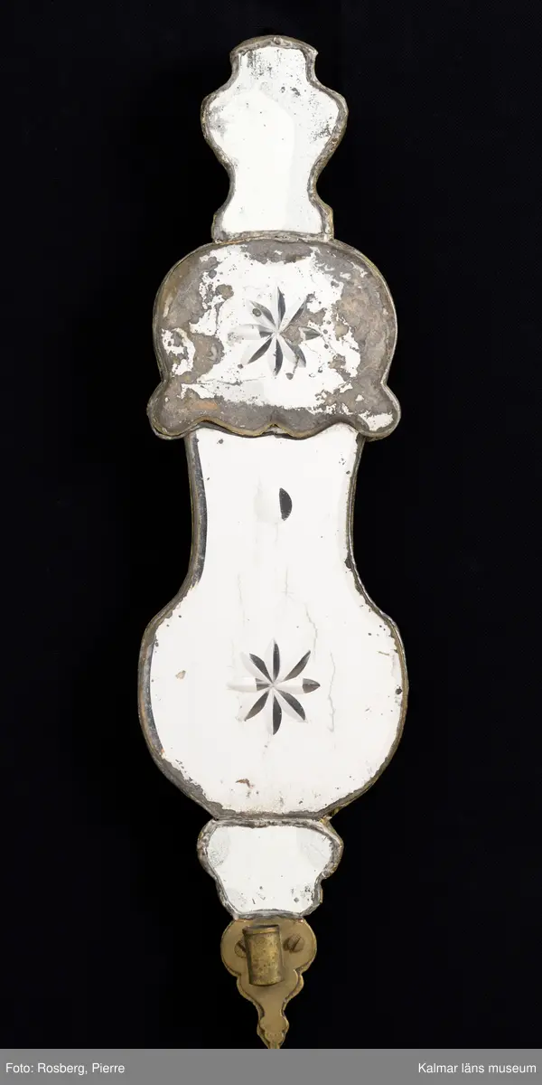 KLM 7184. Lampett, spegellampett. Av trä, bly och spegelglas. Flera utbuktningar, fiolformad, kurvig i flera avdelningar. Väggskivan med profilerad kant, belagd med fyra spegelskivor, fästa med blyram. Väggskivan/bakstycket av trä med förgyllda kanter. Två av spegelskivorna med 8-uddiga stjärnor och en enkel rund figur. Avsedd för en ljushållare, infästning av metall.