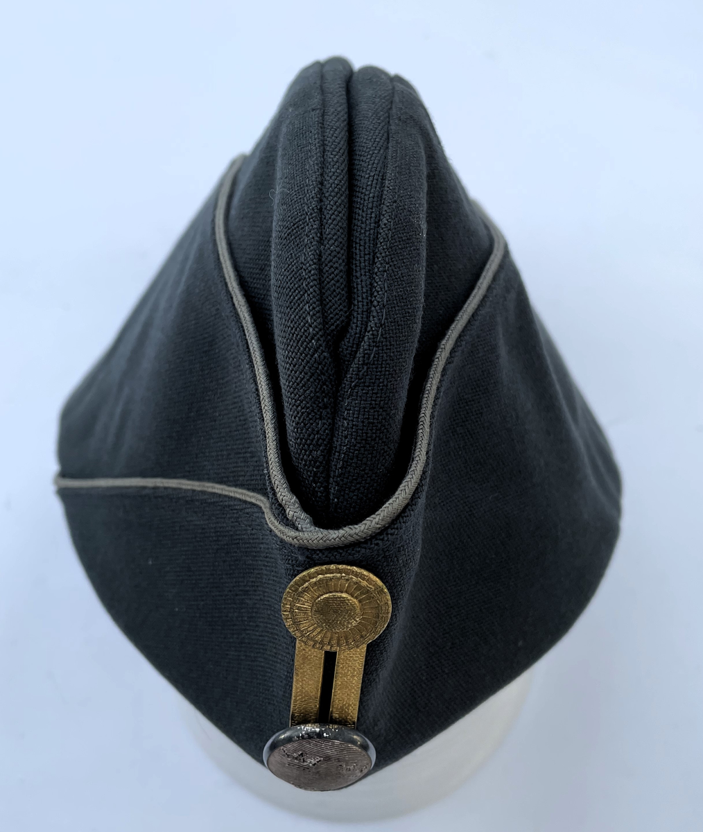 Stålgrå vintermössa m/1960 med gråblå passpoal. Agraff i guld.  Knapp för underofficer m/1865-1960. Vintermössa m/1960 är i samma utförande som Båtmössa m/1960, men har öronskydd och vaddstickat foder.