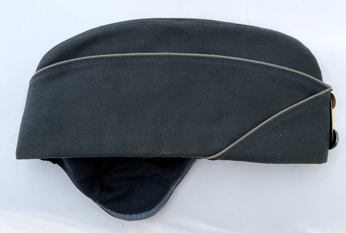 Stålgrå vintermössa m/1960 med gråblå passpoal. Agraff i guld.  Knapp för underofficer m/1865-1960. Vintermössa m/1960 är i samma utförande som Båtmössa m/1960, men har öronskydd och vaddstickat foder.