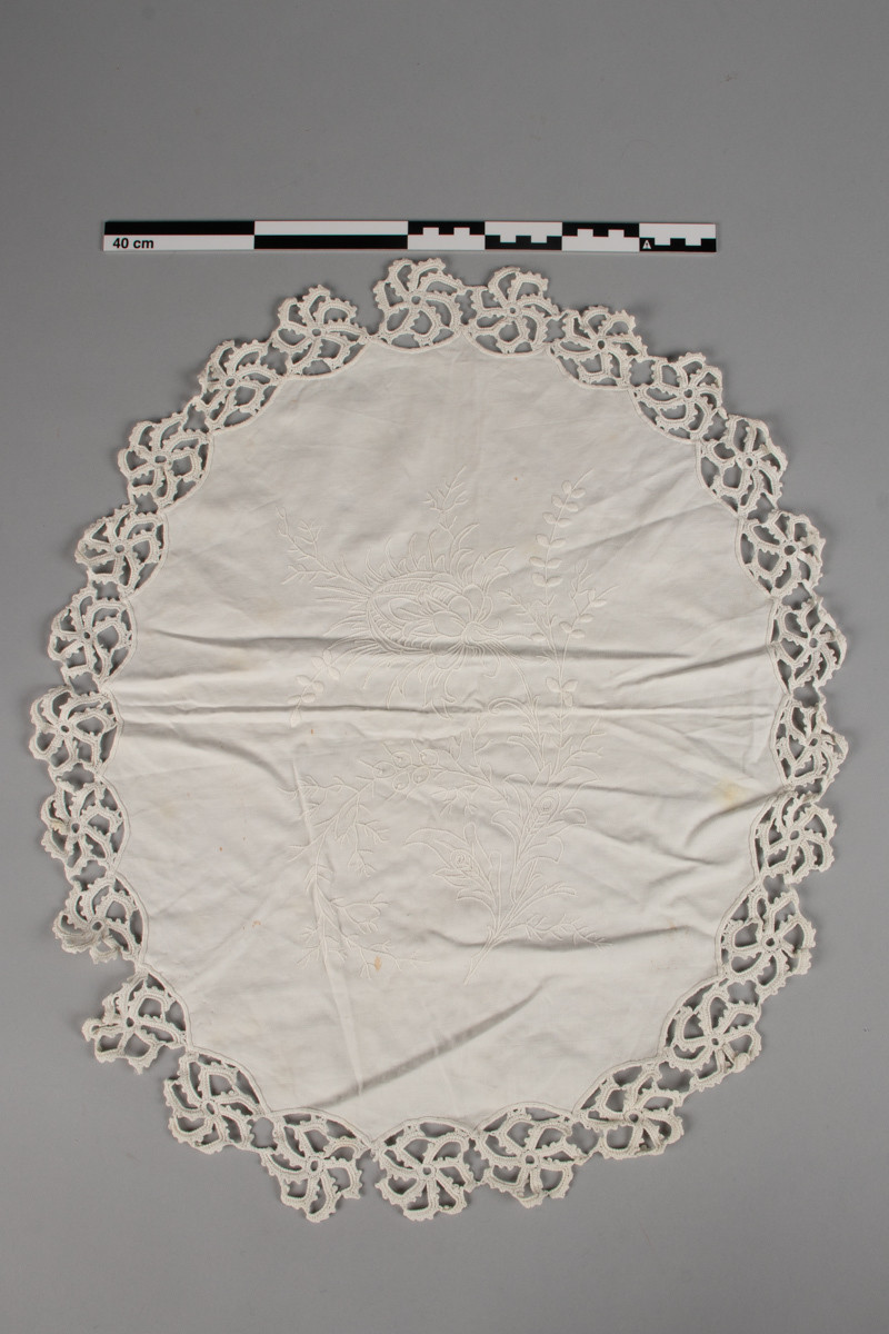 Oval, nesten rund duk med brodert blomstermotiv, hvit tråd på hvit tekstil. Heklet blondekant. Noen flekker.