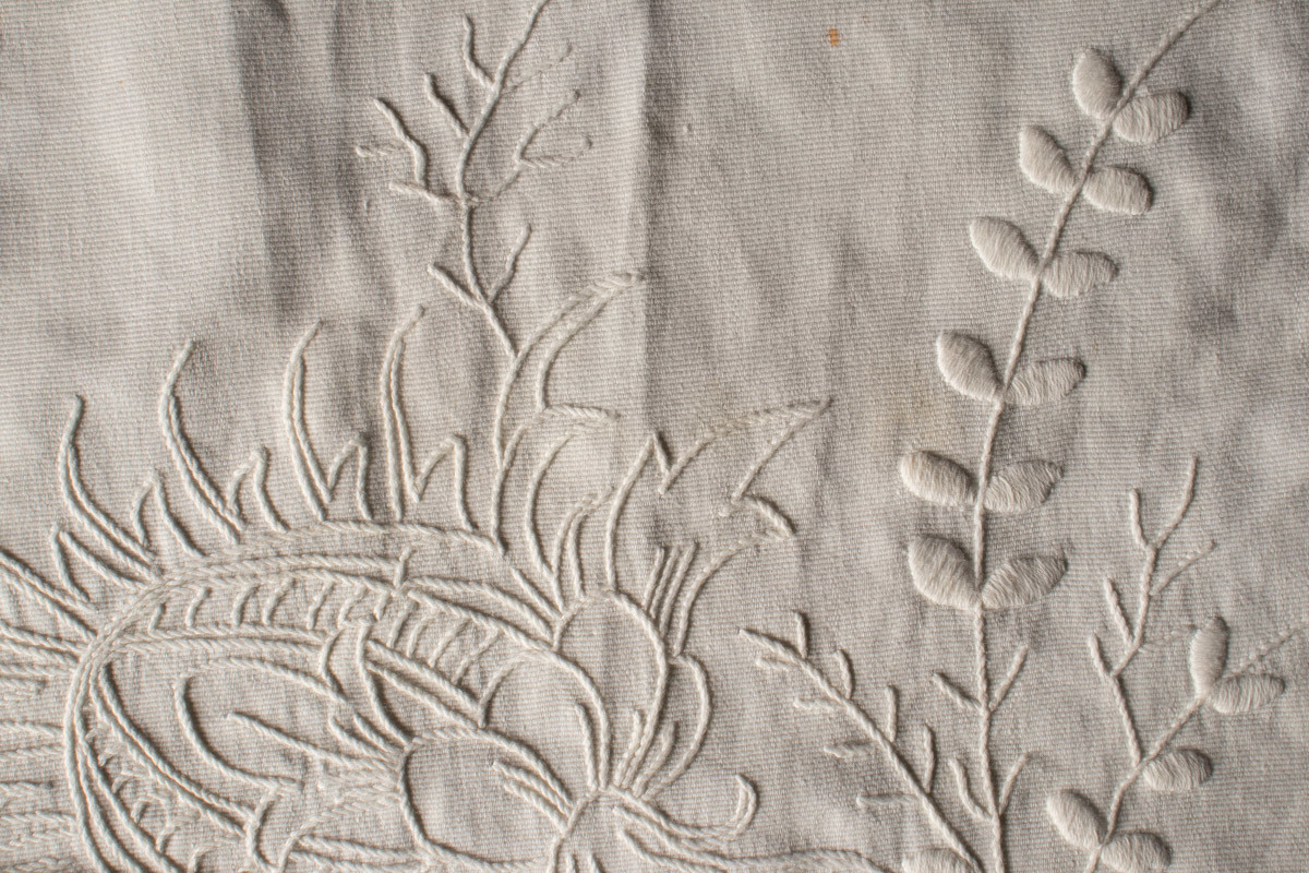 Oval, nesten rund duk med brodert blomstermotiv, hvit tråd på hvit tekstil. Heklet blondekant. Noen flekker.