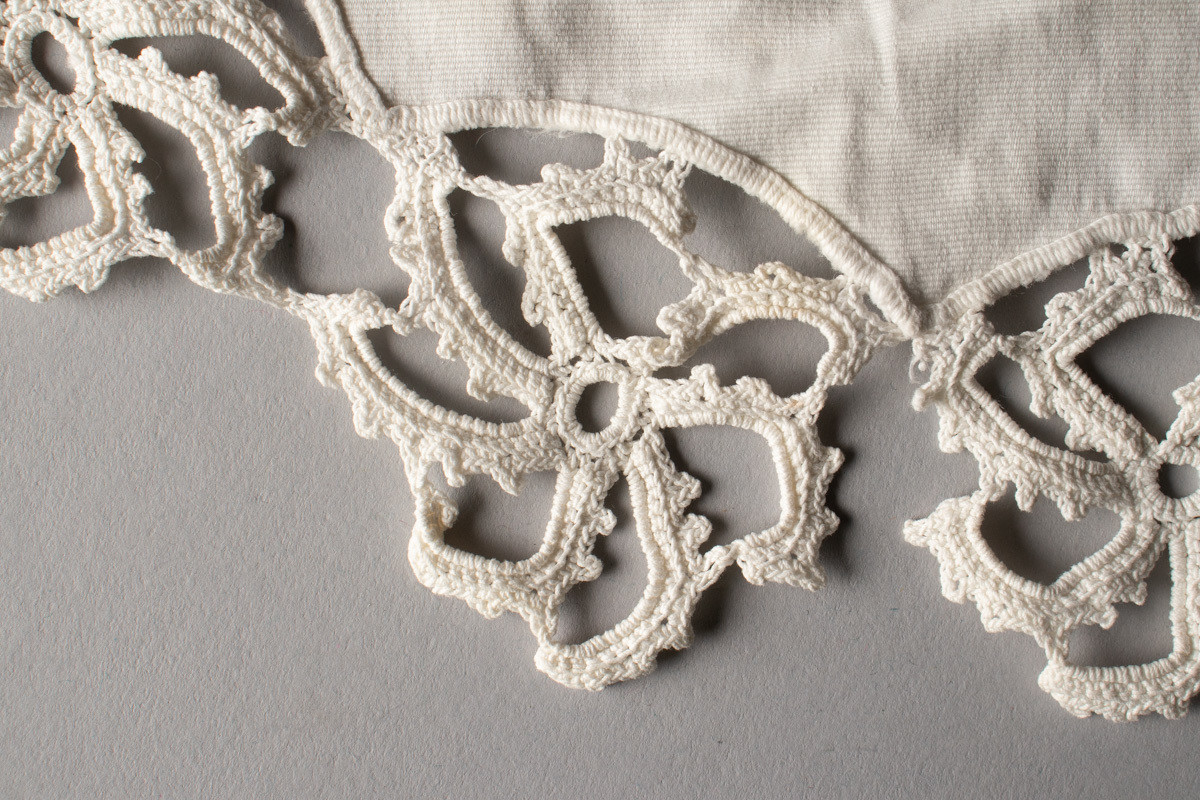 Oval, nesten rund duk med brodert blomstermotiv, hvit tråd på hvit tekstil. Heklet blondekant. Noen flekker.
