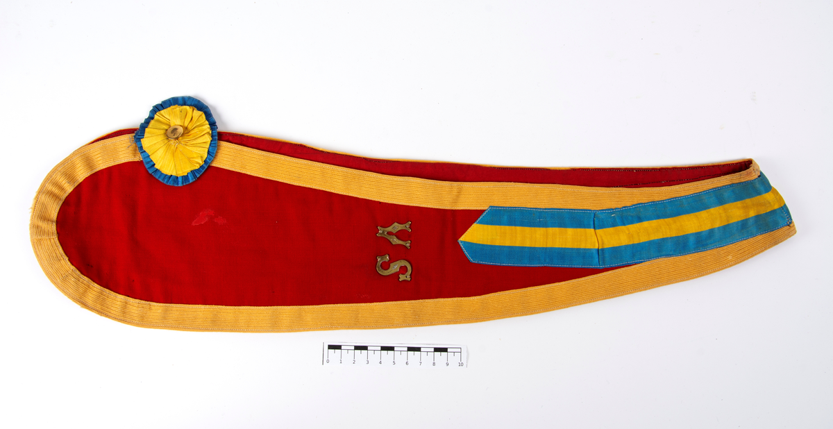 Logeregalie. Bröstpåhäng. Band i rött tyg med droppformade ändar, kantat med gult bårdband. Hålls samman vid bröstet med en tygblomma i gult och blått med gul metallknapp. Vid halspartiet ett påsytt sidenband med de svenska nationalfärgerna. Märkt: "V S". Vice Sekreterare."