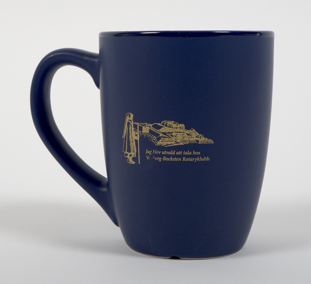 Kaffemugg av blåglaserat stengods. Rak, cylindrisk form, med öra. Två grafiska tryck i guldfärg på vardera sida av muggen. På ena sidan: Rotary international-logotyp. På motsatt sida: Vy över Varbergs fästning med Bockstensmannen i förgrunden, samt texten "Jag blev utvald att tala hos Varberg-Bocksten Rotaryklubb."