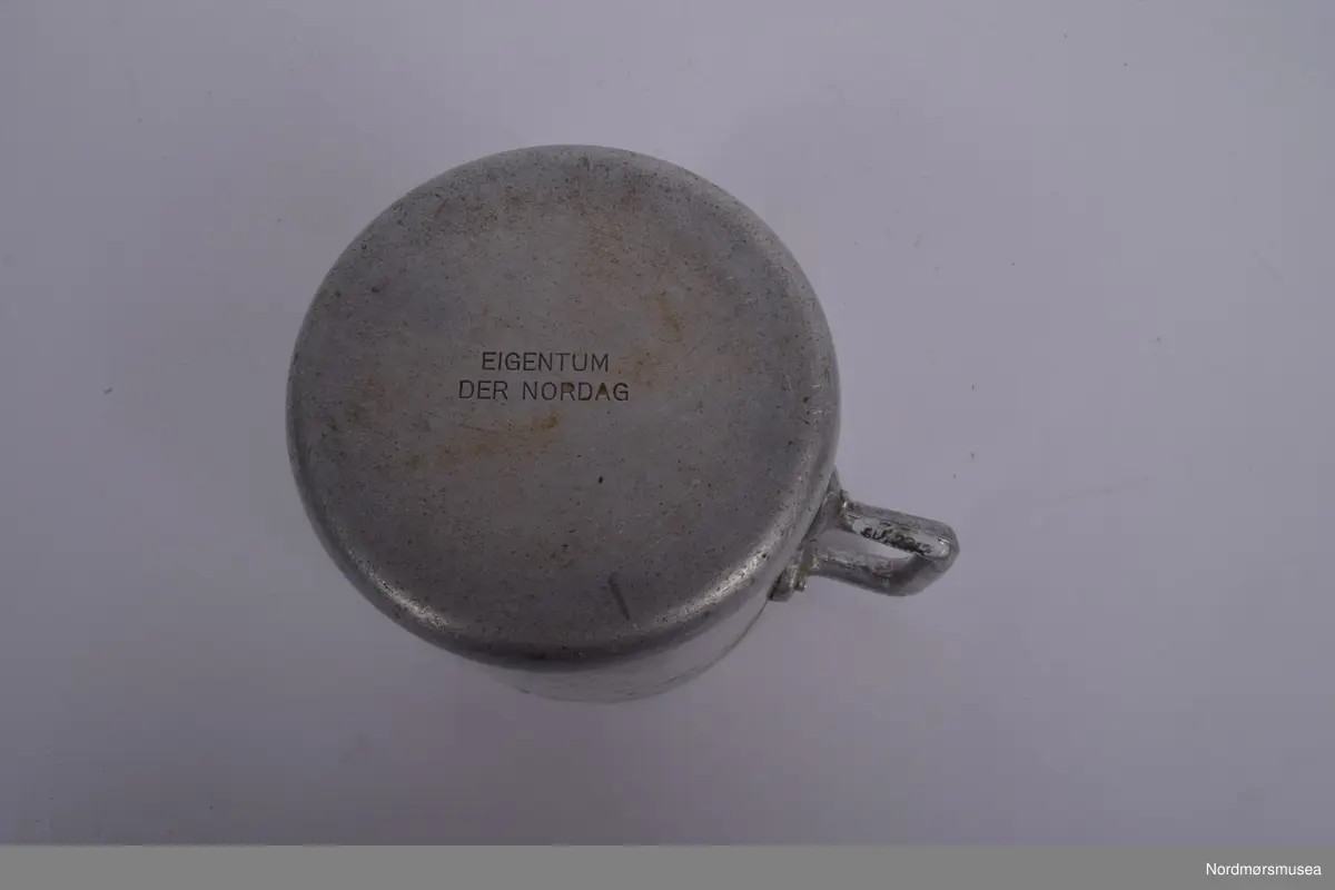 Kopp i alumium med stempel under. Hanken festet med to alumiumsnagler øverst, en nederst.