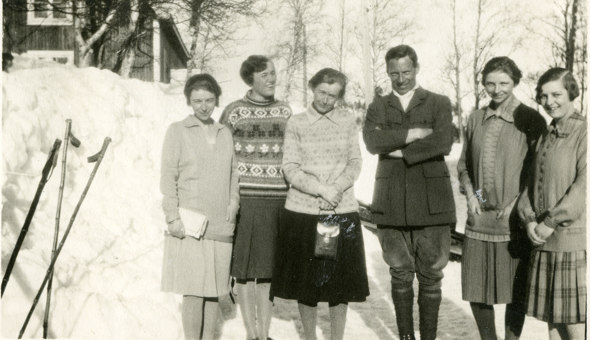 Gruppe av 5 damer i skjørt og en mann foran et hus, det er vinter.
