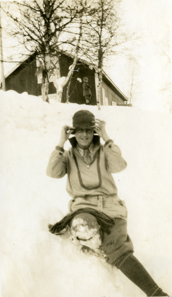 Turistme. Dame sitter i snøen , hus/hytte i bakgrunn på Gjeilo.
