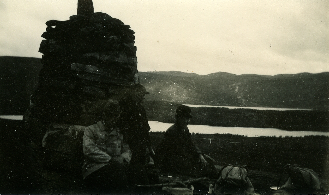 Kveldsstemning på Skøtreberget i Børtnesmarka. Langevatn og Buvatn i bakgrunnen med Hallingnatten i det fjerne. Olav Sveinsson, f. Høllo, John Lunde Sveinsson og Caroline Lunde Sveinsson. Skøtreberget var et sted med god utsikt over beiteområdene i Børtnesmarka og seterfolket dro ofte hit for å skøtre etter beitefeet. Når det stundet mot helg dro de hit for å møte karene på veg innover etter ukedager med slåttearbeid hjemme på gården nede i bygda.
