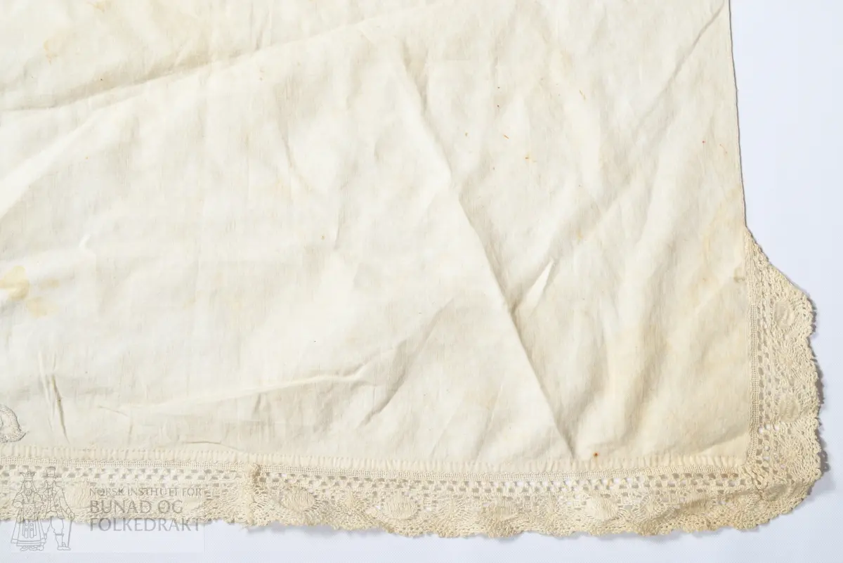 Hatteklede, skaut.
Av tynn, fin kvit bomullscambrik.
Kvadratisk, der eine snippen er brodert med plattsaum i fleire falma fargar, lys beige, kvitgrå, 
lys blå blank lintråd, og raud lettfalma bomullstråd.
Brodert AJD 1851 i ei midtrose. 
Påsydd maskinvirka blonde av kvit lin langs kanten av snippen.