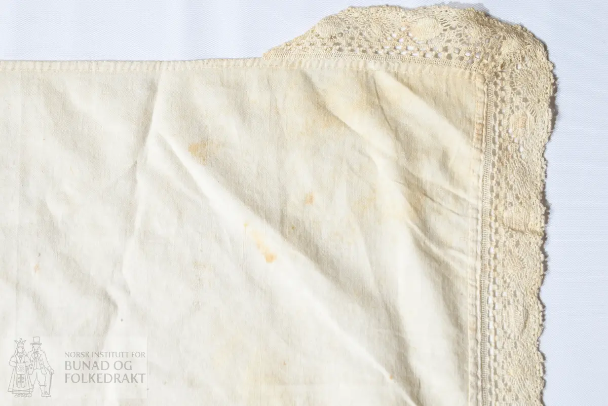 Hatteklede, skaut.
Av tynn, fin kvit bomullscambrik.
Kvadratisk, der eine snippen er brodert med plattsaum i fleire falma fargar, lys beige, kvitgrå, 
lys blå blank lintråd, og raud lettfalma bomullstråd.
Brodert AJD 1851 i ei midtrose. 
Påsydd maskinvirka blonde av kvit lin langs kanten av snippen.