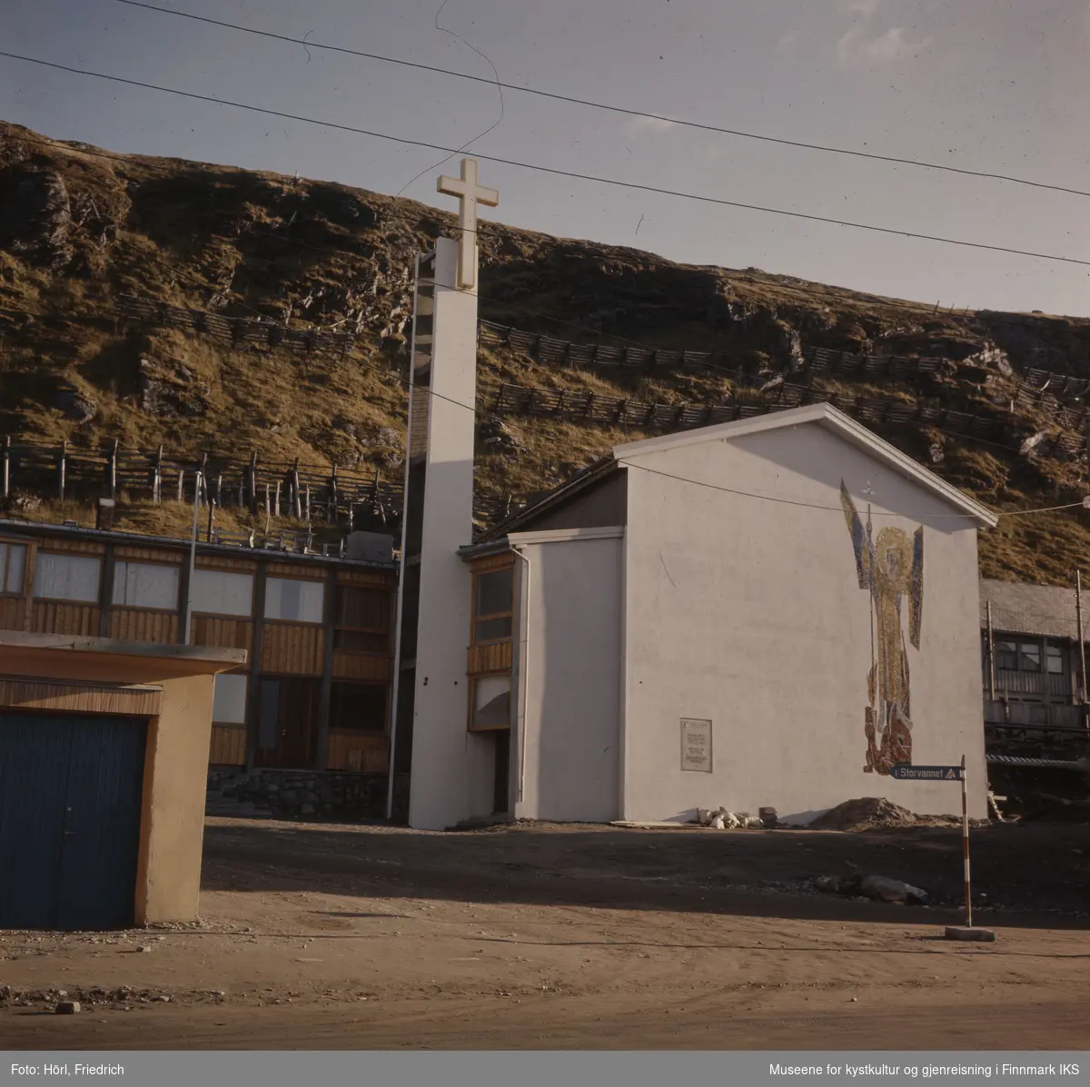 Den nybygde katolske kirke St. Mikael i Hammerfest står ferdig en solskinnsdag i 1958. Til venstre for tårnet ser man et tilbygg. Kirka er fotografert fra Strandgata og man ser Salen fjellet i bakgrunnen. På kirkens yttervegg ser man tydelig den store mosaikken som viser St. Mikael som er skapt av den tyske billedkunstneren Karl Manninger.