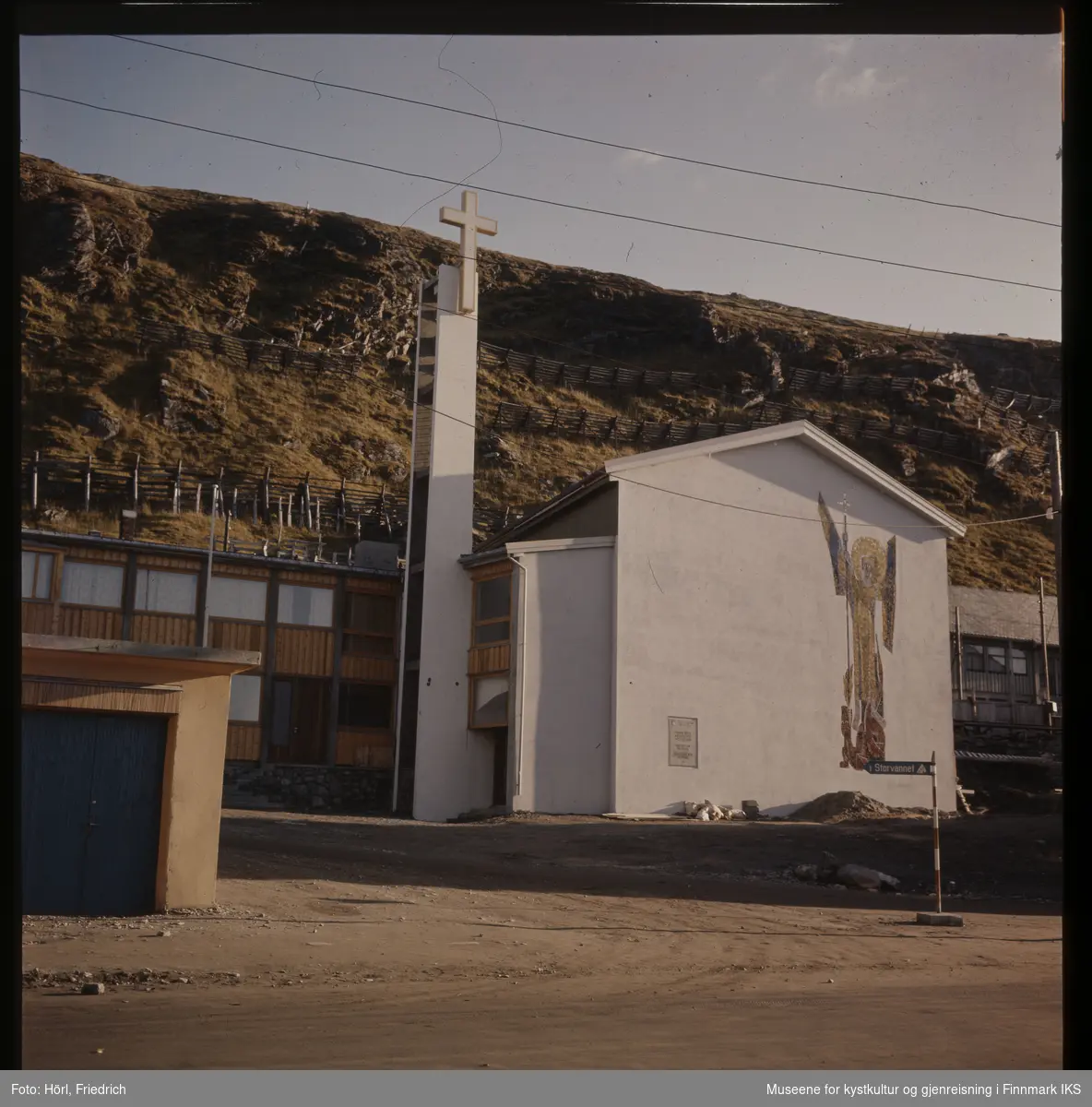 Den nybygde katolske kirke St. Mikael i Hammerfest står ferdig en solskinnsdag i 1958. Til venstre for tårnet ser man et tilbygg. Kirka er fotografert fra Strandgata og man ser Salen fjellet i bakgrunnen. På kirkens yttervegg ser man tydelig den store mosaikken som viser St. Mikael som er skapt av den tyske billedkunstneren Karl Manninger.