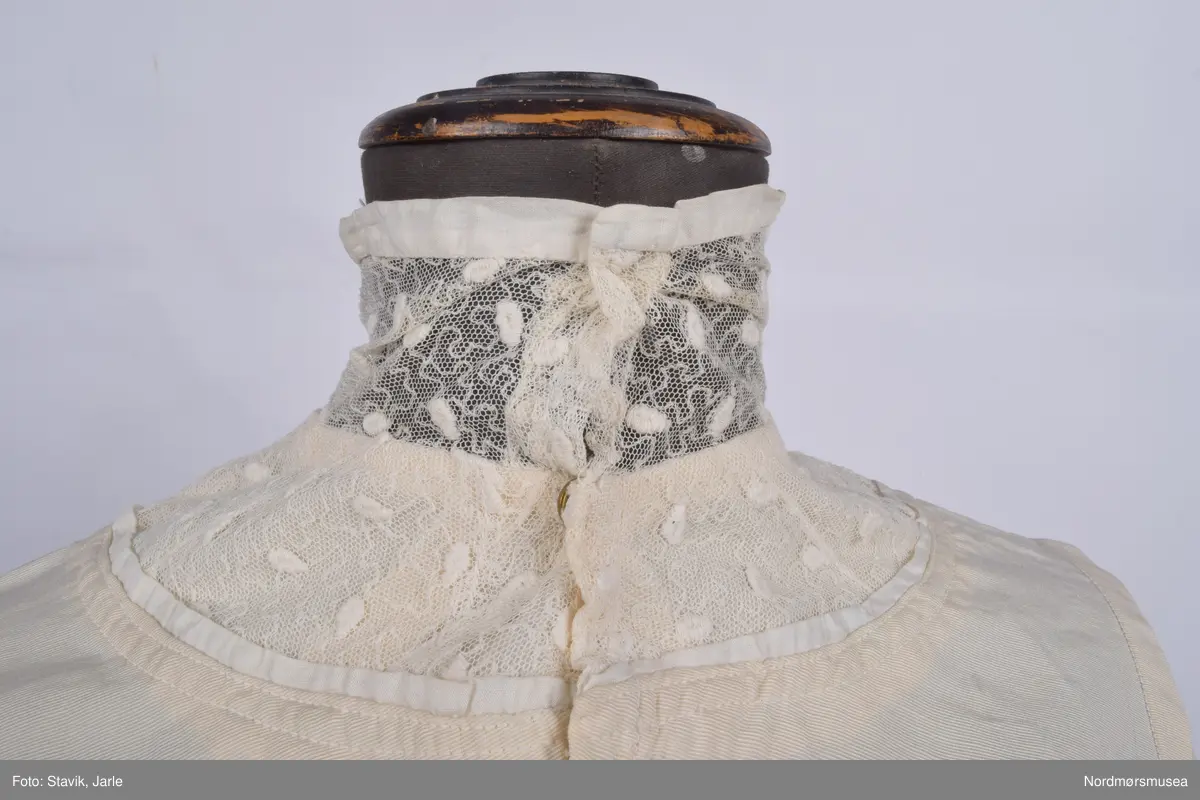 Skjorte i naturhvit lin med påsydd blondekrage. kragestoffet er påsydd slik at omtrent halvparten går nedenfor kragen, mens resten danner kragen. Trykknapper i ryggen. Ermene er laget for å brettes lett opp i enden. I front er stofet brett og deretter sydd i en stripe som går fra skulderen ned til livet. Denne stripen fortsetter på baksiden på samme vis. To ekstra striper i front. Tre stoffkledde pynteknapper er plassert øverst ved hver stripe. Bortsett fra enkelte påsydde stoffbiter slik som innlegg under armene ser skjorten ut til å være laget av ett stoffstykke tilklipt i form.