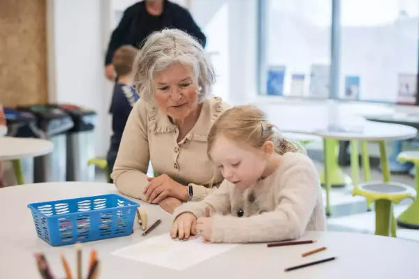 En eldre dame sitter til venstre i bildet ved siden av en liten jente. De er konsentrert over en tegning som jenta holder på med.