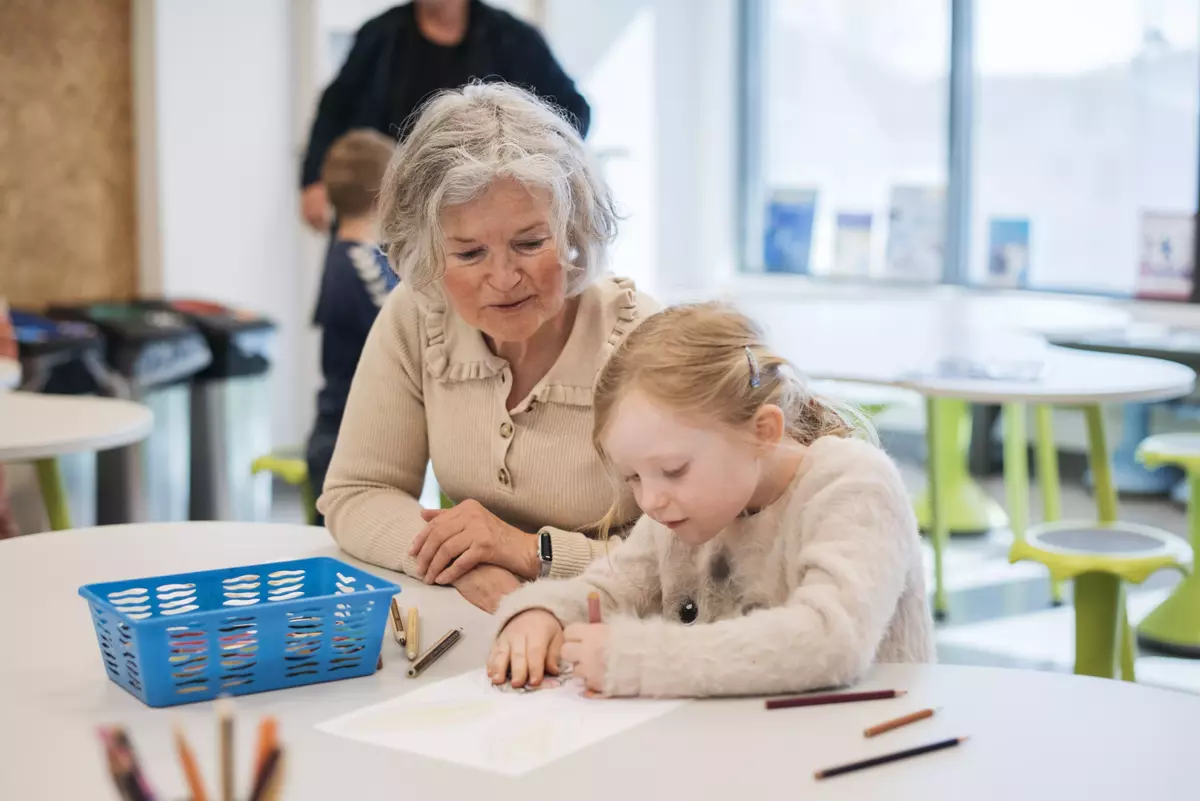 En eldre dame sitter til venstre i bildet ved siden av en liten jente. De er konsentrert over en tegning som jenta holder på med.