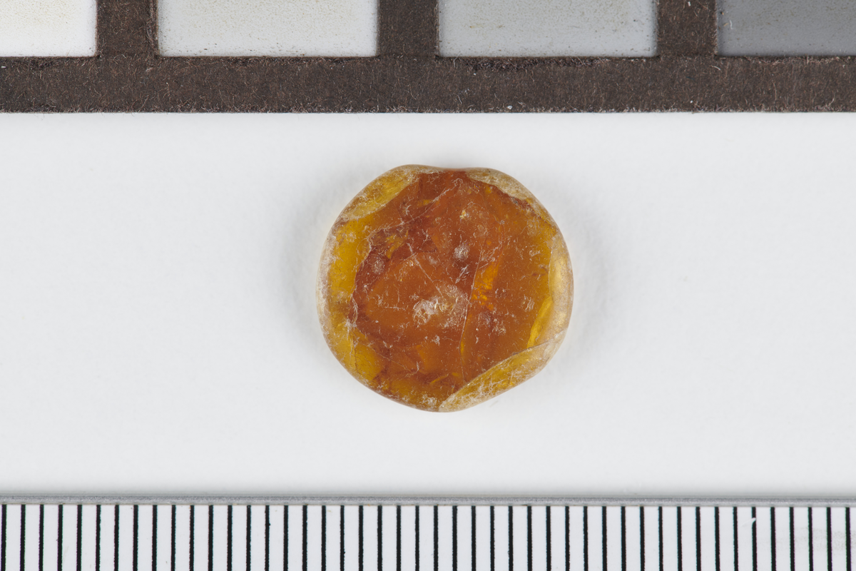 Flat, tilnærmet rund skive av rav. Mål: Diam: 1,3 cm. T: 0,3 cm. Vekt: 0,9 gram.