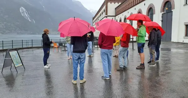 Bilete av sommarvert i aksjon utforbi kraftstasjonen, den regnar og det står fleire personer med paraply å høyre på kva verten har å fortelje