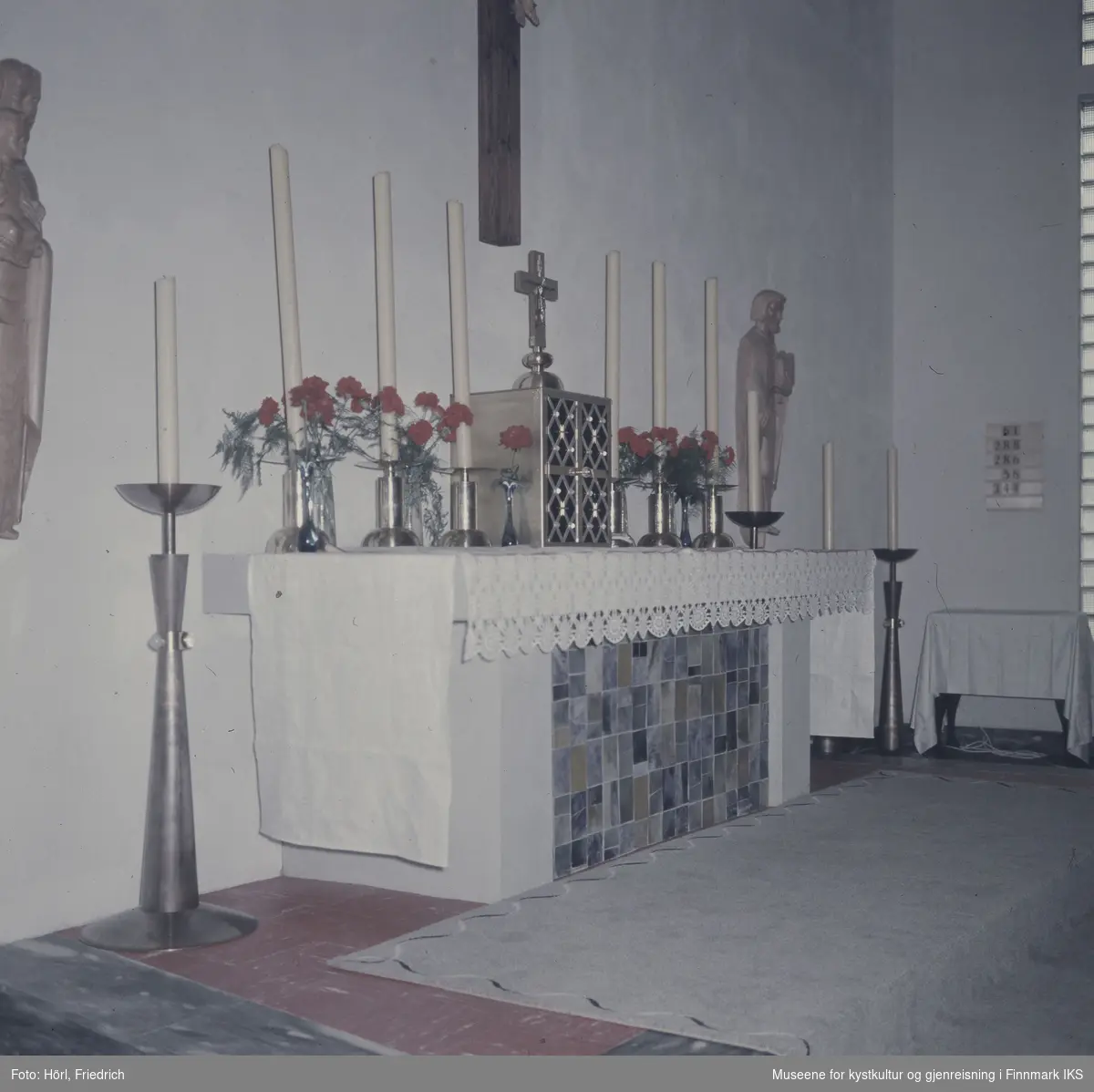 Interiøret i Den katolske kirke St. Mikael i Hammerfest i 1958. Bildet viser alteret med forsoningskorset ved veggen over det og treskulpturer til venstre og høyre for det. På alteret ligger det et hvit duk, i midten er alterskapet plassert og stearinlys og røde nelliker står ved siden av det. I tillegg er det lysestaker med stearinlys plassert på gulvet ved siden av alteret.