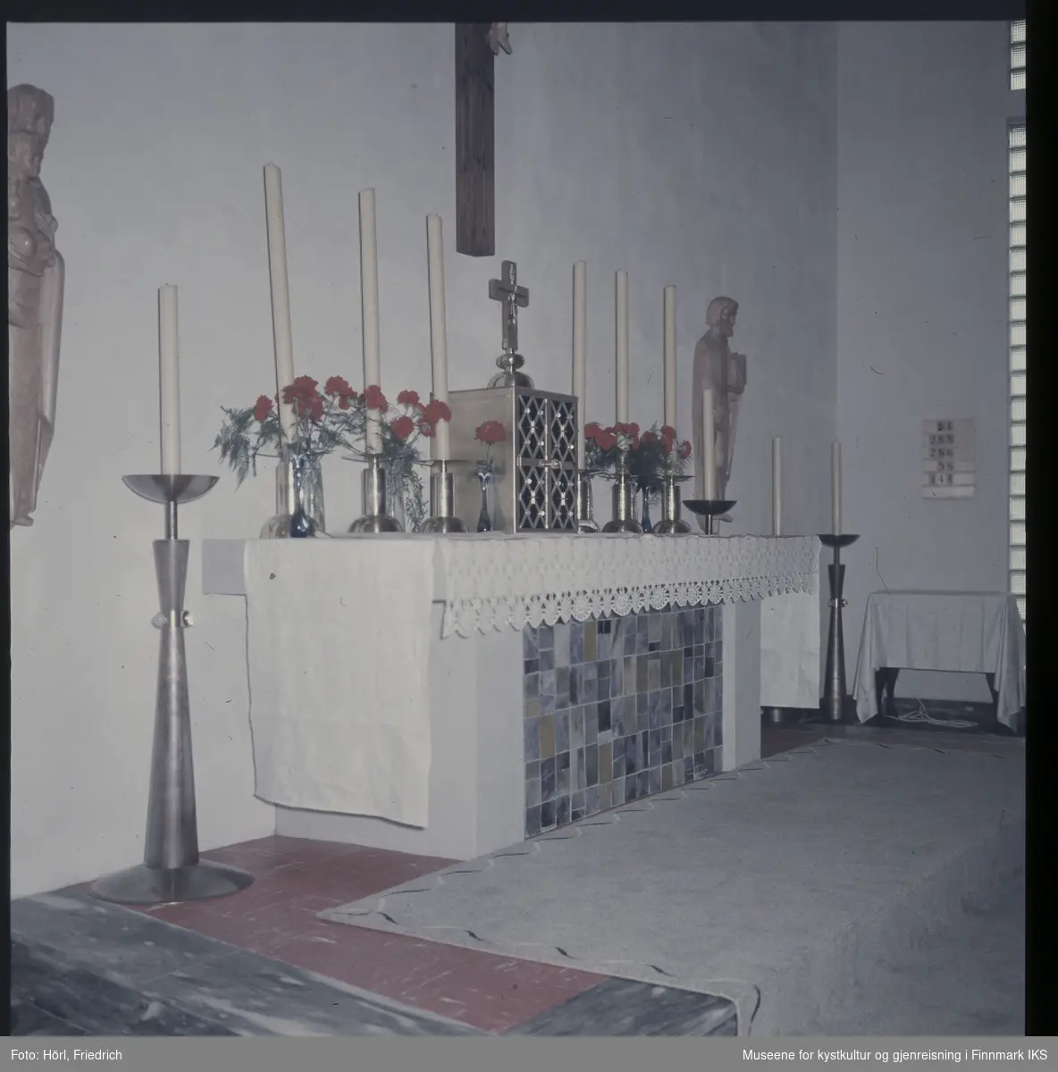 Interiøret i Den katolske kirke St. Mikael i Hammerfest i 1958. Bildet viser alteret med forsoningskorset ved veggen over det og treskulpturer til venstre og høyre for det. På alteret ligger det et hvit duk, i midten er alterskapet plassert og stearinlys og røde nelliker står ved siden av det. I tillegg er det lysestaker med stearinlys plassert på gulvet ved siden av alteret.