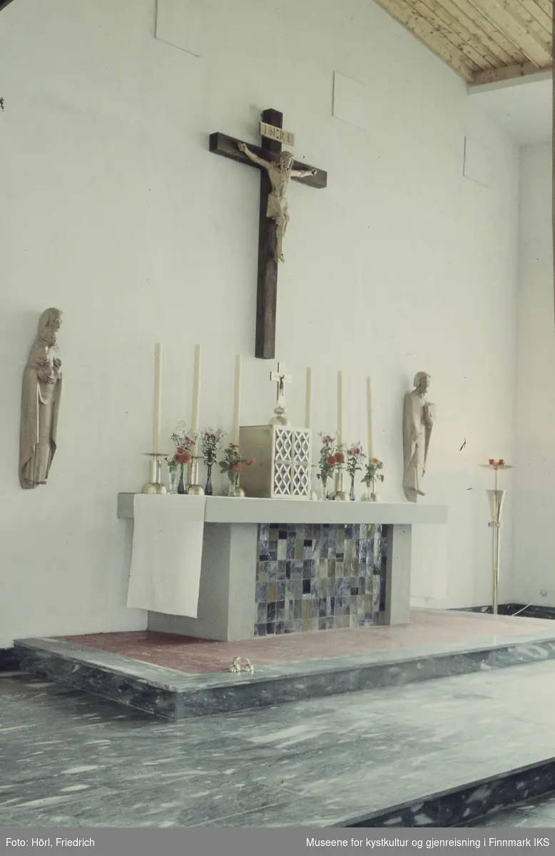 Bildet viser interiøret til Den katolske kirke St. Mikael i Hammerfest. I midten ser vi alteret som er pyntet med duk, stearinlys og blomsterbuketter. I midten på alteret står tabernakelet som tjener til oppbevaring av hostiene. Oppå den står et kors. Ovenfor alteret henger det en fremstilling av Jesus på korset, skjært i tre. Ved siden av alteret er det to ytterligere treskulpturer på veggen. Til venstre for alteret står en gulvlampe. På gulvet ligger det en bjelle.