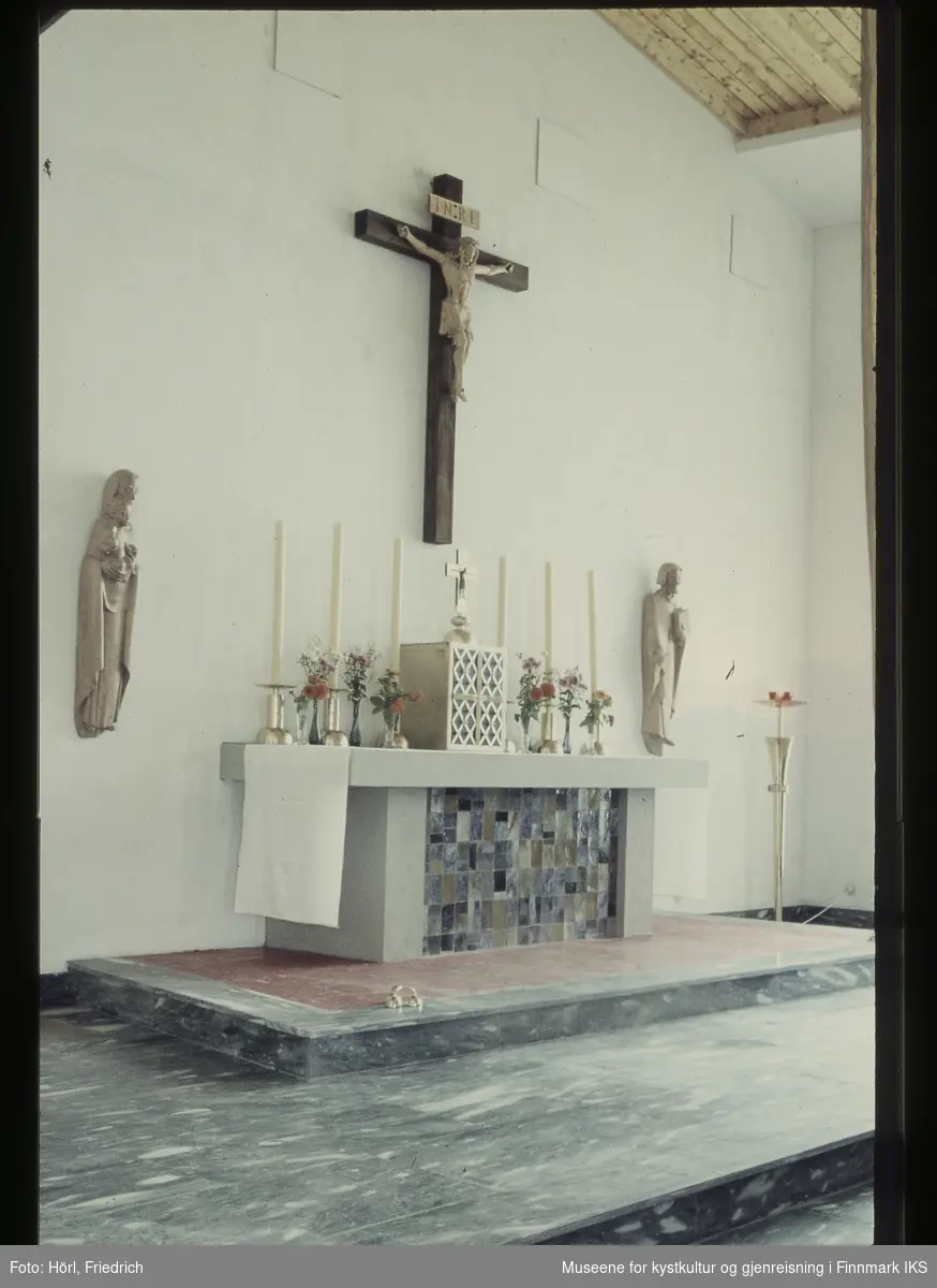 Bildet viser interiøret til Den katolske kirke St. Mikael i Hammerfest. I midten ser vi alteret som er pyntet med duk, stearinlys og blomsterbuketter. I midten på alteret står tabernakelet som tjener til oppbevaring av hostiene. Oppå den står et kors. Ovenfor alteret henger det en fremstilling av Jesus på korset, skjært i tre. Ved siden av alteret er det to ytterligere treskulpturer på veggen. Til venstre for alteret står en gulvlampe. På gulvet ligger det en bjelle.