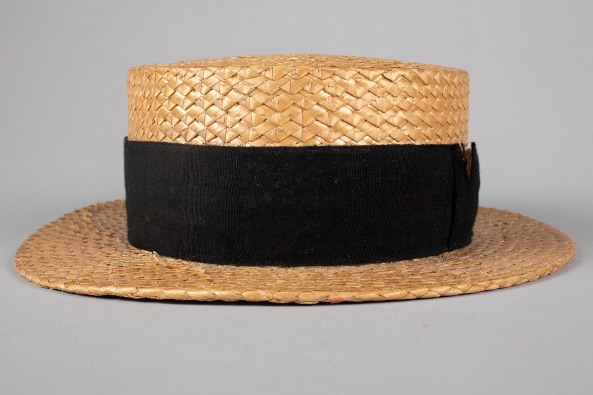 Stråhatten har en flat rett pull og bred flat brem. Svart bånd rundt pullen med fast sydd sløyfe. Svettebånd i lær og netting fôr. I bunnen er det festet et tøystykke med innlegg og logo. Bremen har fått en knekk og hattebåndet har en rift. Halmen er flettet med taggemønster. 