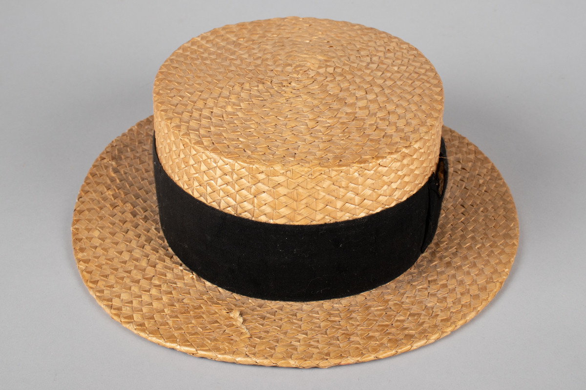 Stråhatten har en flat rett pull og bred flat brem. Svart bånd rundt pullen med fast sydd sløyfe. Svettebånd i lær og netting fôr. I bunnen er det festet et tøystykke med innlegg og logo. Bremen har fått en knekk og hattebåndet har en rift. Halmen er flettet med taggemønster. 