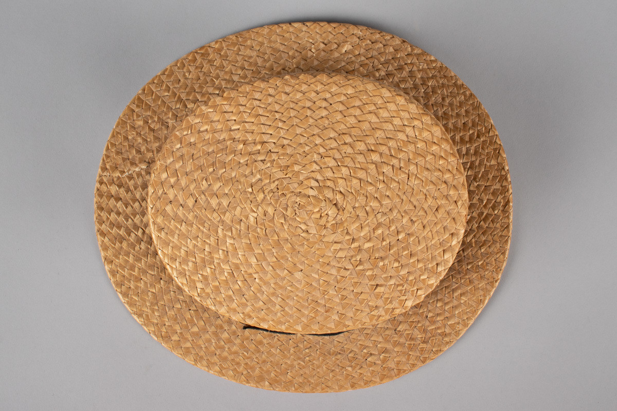 Stråhatten har en flat rett pull og bred flat brem. Svart bånd rundt pullen med fast sydd sløyfe. Svettebånd i lær og netting fôr. I bunnen er det festet et tøystykke med innlegg og logo. Bremen har fått en knekk og hattebåndet har en rift. Halmen er flettet med taggemønster. 