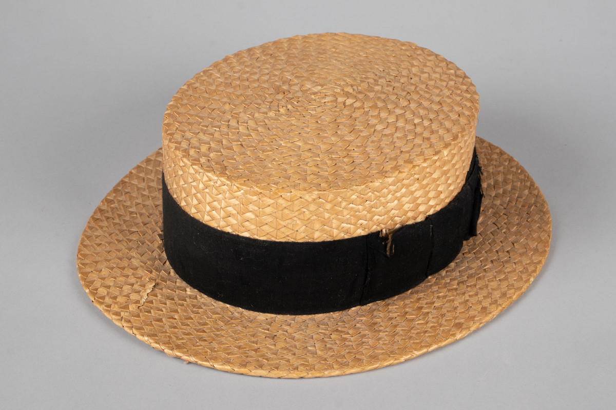 Stråhatten har en flat rett pull og bred flat brem. Svart bånd rundt pullen med fast sydd sløyfe. Svettebånd i lær og netting fôr. I bunnen er det festet et tøystykke med innlegg og logo. Bremen har fått en knekk og hattebåndet har en rift. Halmen er flettet med taggemønster. 