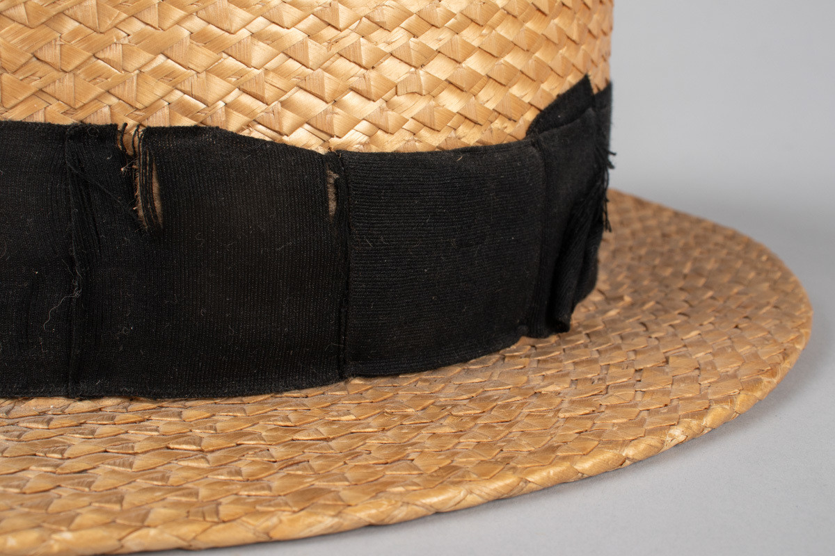 Stråhatten har en flat rett pull og bred flat brem. Svart bånd rundt pullen med fast sydd sløyfe. Svettebånd i lær og netting fôr. I bunnen er det festet et tøystykke med innlegg og logo. Bremen har fått en knekk og hattebåndet har en rift. Halmen er flettet med taggemønster. 
